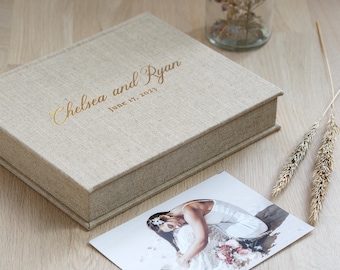 May include: A rectangular beige linen-covered photo album box with the gold foil text "Chelsea and Ryan" and "June 17, 2023" printed on the lid. A rectangular photograph showing a person in a white dress is placed next to the box.