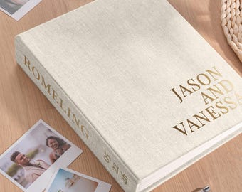 May include: A cream-colored linen wedding photo album with gold lettering that reads "Jason and Vanessa" on the front and "Romeling" on the spine. The album is surrounded by printed photos and a pen.