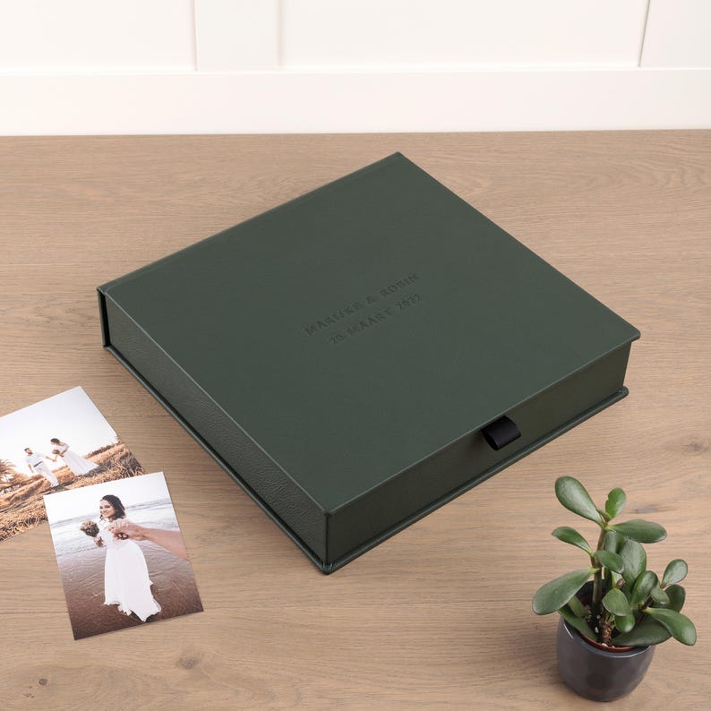 May include: A dark green, square box with a black ribbon pull, likely for storage or presentation. The box has embossed text on the top. Two photographs and a small potted plant are next to the box on a wooden surface.