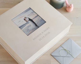 Op de afbeelding: Een vierkant, crèmekleurig fotoalbum met een gestructureerde kaft en een centraal vierkant venster met een foto van een koppel dat elkaar omhelst op een strand. De kaft is voorzien van de tekst BRENDA & MARK en de datum 01.24.2026.