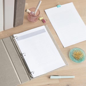May include: A beige three-ring binder containing lined paper with a gray header, a single sheet of lined paper with a blue binder clip, a pink fluted pen holder with pens, several colorful binder clips, a glass dish of gold paper clips, and a pen.