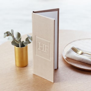 Könnte beinhalten: Eine hohe, beige, texturierte Menükarte mit einem geprägten quadratischen Rahmen, der die Buchstaben HF und das Wort ADVISORS enthält. Die Karte steht aufrecht, zeigt weiße Innenseiten und ist mit einem stabilen, buchähnlichen Rücken versehen.
