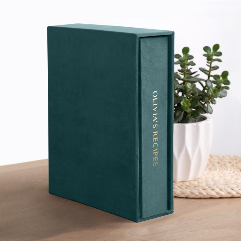 May include: A dark teal velvet-covered box with a rectangular shape. The spine features the gold-colored text OLIVIA'S RECIPES printed vertically. The box has a smooth, matte texture and a clean, structured design with sharp, defined edges.