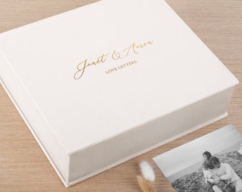 May include: A square, off-white keepsake box with a hinged lid. The top features gold foil text that reads "Janet & Aaron" in a cursive script above the words "LOVE LETTERS" in a clean, sans-serif font. A black and white photograph sits nearby.