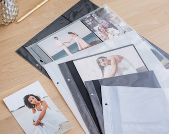 May include: A set of clear plastic photo album refill pages with reinforced black and gray edges featuring multiple pockets for photographs. The pages are arranged in a stack, with several printed photographs visible inside the clear sleeves.