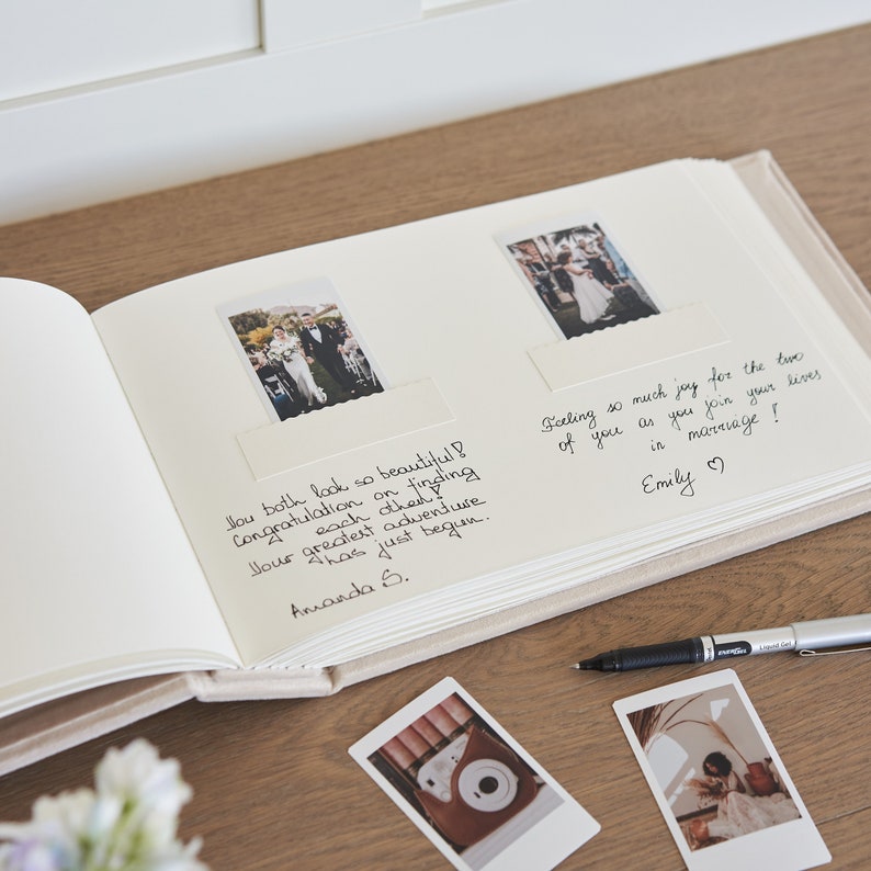 Puede incluir: Un &aacute;lbum de fotos de boda beige abierto, con dos fotos y mensajes manuscritos. Un bol&iacute;grafo negro y dos fotos adicionales est&aacute;n sobre la superficie de madera. El &aacute;lbum tiene una cubierta de pl&aacute;stico transparente.