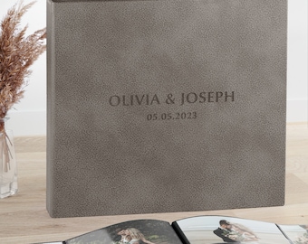 May include: A square, textured gray photo album with a leather-like finish. The front cover is embossed with the text OLIVIA & JOSEPH and the date 05.05.2023. The album is displayed upright, with an open photo album showing wedding pictures below.