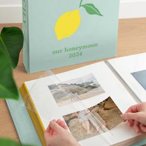 Op de afbeelding: Een lichtgroen fotoalbum met een gele citroenillustratie en de tekst "our honeymoon 2024" op de kaft. Het album is geopend en toont witte pagina's met doorzichtige plastic hoezen waarin foto's van een koppel op een strand zitten.