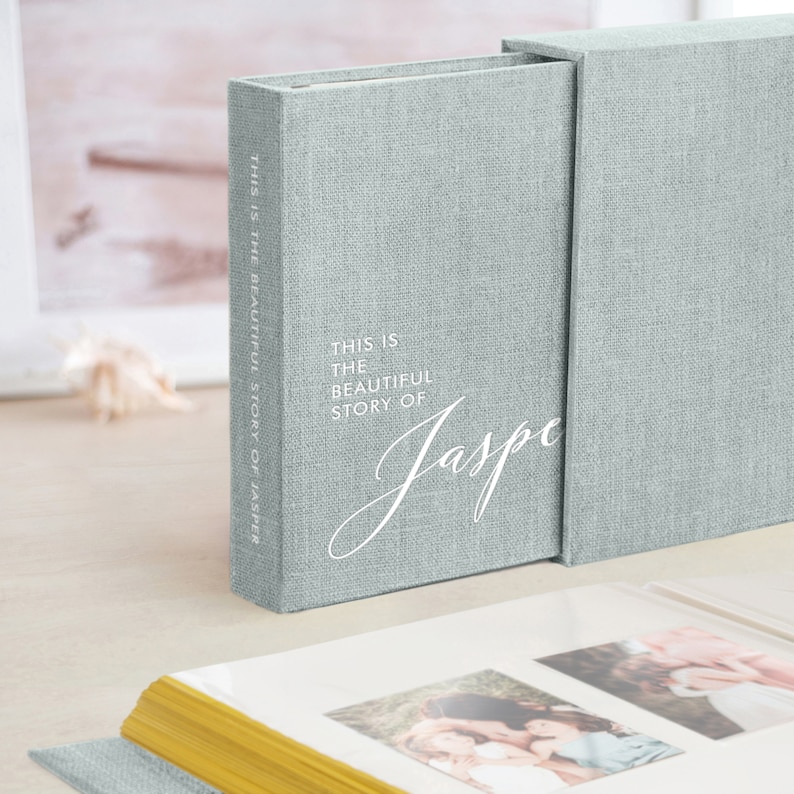 May include: A light blue linen photo album with a matching slipcase. The album cover features the text "THIS IS THE BEAUTIFUL STORY OF Jasper" in white script. The album is open, revealing photos.
