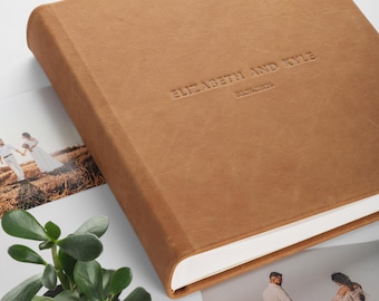May include: A square photo album with a tan leather cover featuring the embossed text ELIZABETH AND KYLE and the date 02.25.2025. The album has white pages visible along the edge and is positioned near two photographs and a small green plant.