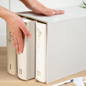 May include: A white storage box with three decorative books. The books are off-white and beige with gold lettering that reads "Harris" and Roman numerals. The box is being opened by a person's hand.
