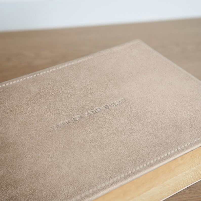 May include: A close-up of a beige leather-bound book with the embossed text "PATRICK AND HELEN". The book has a stitched border and rests on a light-colored wooden surface. The cover has a textured appearance.