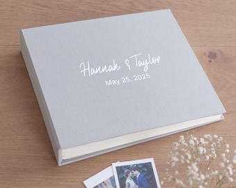 May include: A light gray square photo album with a hard cover featuring the white printed text Hannah & Taylor and the date May 25, 2025. The album has a clean, solid-colored exterior and is positioned next to two small rectangular photographs.