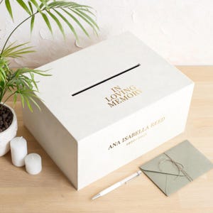 May include: A cream-coloured box with a slot, gold text reading "IN LOVING MEMORY" and the name "ANA ISABELLA REED 1950-2023". A pen and envelope are next to the box. Candles and a plant are in the background.
