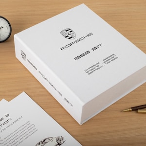 Peut inclure: Une boîte rectangulaire blanche arborant le logo Porsche noir et le texte 1969 911T. La surface supérieure affiche les numéros de châssis, moteur et boîte de vitesses. Le côté de la boîte répète 1969 PORSCHE 911T. Un document partiel est visible.
