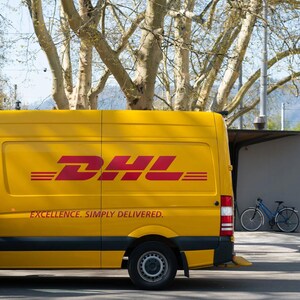 May include: A yellow delivery van with the DHL logo and the slogan "Excellence. Simply delivered." on the side. The van is parked in front of a building with trees in the background.