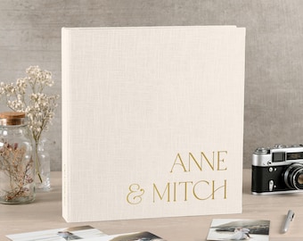 Könnte beinhalten: Ein quadratisches Fotoalbum mit einem cremefarbenen Leineneinband und dem goldgeprägten Text ANNE & MITCH. Das Album steht neben einem Glas mit Trockenblumen, einer Vintage-Kamera, einem silbernen Stift und zwei losen Fotografien.