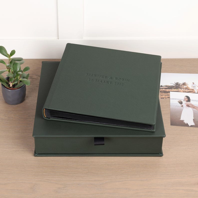 May include: A dark green photo album with the embossed text "MARTINE & ROBIN 10 MAART 2022" on the cover, resting on a matching box. A small potted plant and a photograph are visible in the background. The album has a gold-colored spine.