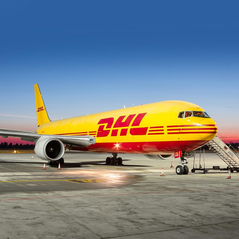 May include: A yellow and red DHL Boeing 767-300F cargo plane on the tarmac at an airport. The plane is parked with its nose facing the camera. The plane has a DHL logo on the side.