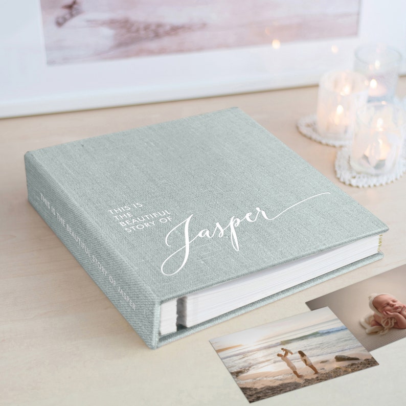 May include: A light green linen-covered photo album with the name "Jasper" in white script. The album's spine reads "THIS IS THE BEAUTIFUL STORY OF JASPER". Two photos are visible, along with lit candles in glass holders.