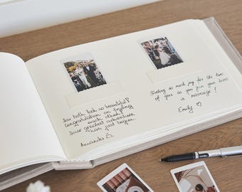 May include: An open wedding photo album with two photos and handwritten messages. The album is on a wooden surface, with a pen and two additional photos nearby. The text reads: "How both look so beautiful! Congratulations on finding each other! Your greatest adventure has just begun."