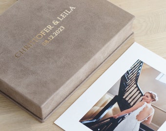 May include: A rectangular taupe suede photo box with gold foil text reading CHRISTOFER & LEILA 08.12.2023. Beside the box lies a white-bordered photograph showing a person in a white backless wedding dress standing on a dark wooden staircase.