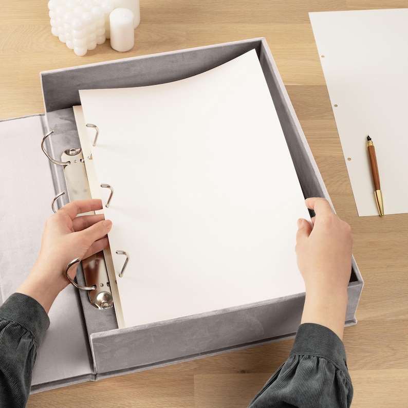 K&ouml;nnte beinhalten: Ein grauer Ordner mit silbernen Ringen, der wei&szlig;es Papier enth&auml;lt. Der Ordner ist ge&ouml;ffnet, mit H&auml;nden, die das Papier halten. Ein Holzstift und ein Blatt Papier mit L&ouml;chern liegen auf der Holzoberfl&auml;che.