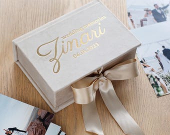 Könnte beinhalten: Eine rechteckige Aufbewahrungsbox aus hellbeigem Samt mit goldfarbenem Folientext, der wedding memories Zinari 06.01.2023 lautet. Die Box ist mit einem breiten, passenden beigen Satinband verschlossen, das vorne rechts zu einer Schleife gebunden ist.