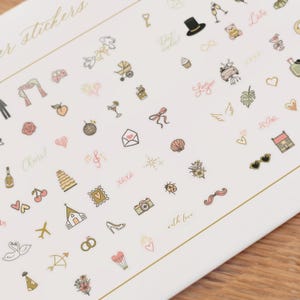 Pode incluir: Uma folha de autocolantes decorativos com v&aacute;rias ilustra&ccedil;&otilde;es em ouro, rosa e preto. Os autocolantes incluem imagens com tema de casamento, como um bolo, an&eacute;is e uma igreja, juntamente com outros desenhos como cora&ccedil;&otilde;es, uma c&acirc;mara e o texto "Cheers!"