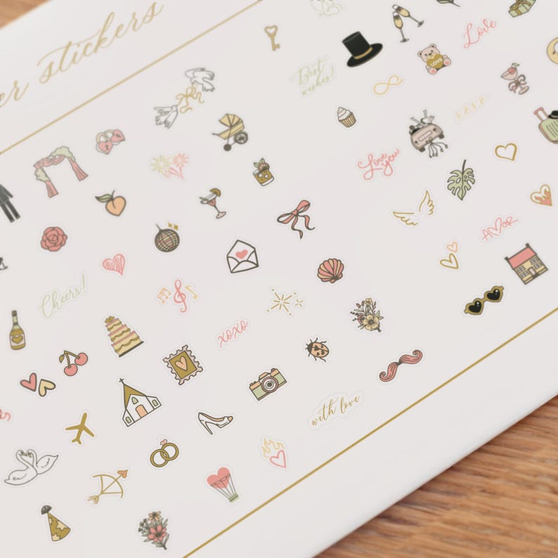 Pode incluir: Uma folha de autocolantes decorativos com v&aacute;rias ilustra&ccedil;&otilde;es em ouro, rosa e preto. Os autocolantes incluem imagens com tema de casamento, como um bolo, an&eacute;is e uma igreja, juntamente com outros desenhos como cora&ccedil;&otilde;es, uma c&acirc;mara e o texto "Cheers!"