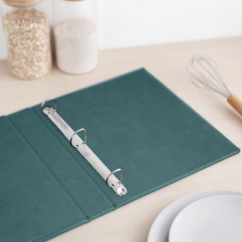 May include: A dark teal three-ring binder featuring a smooth, matte-textured cover and a silver-colored metal mechanism with three circular rings attached to the spine, designed for organizing documents or paper sheets in a professional setting.