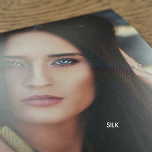 May include: Close-up of a woman's face with blue eyes and long dark hair. The image has a soft, blurred background and the word "SILK" is printed in white at the bottom.