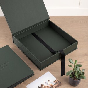 May include: An open, dark green presentation box with a black ribbon, next to a matching album. The box is on a wooden surface, with a small potted plant and a photograph of a couple. The album has text on the cover.