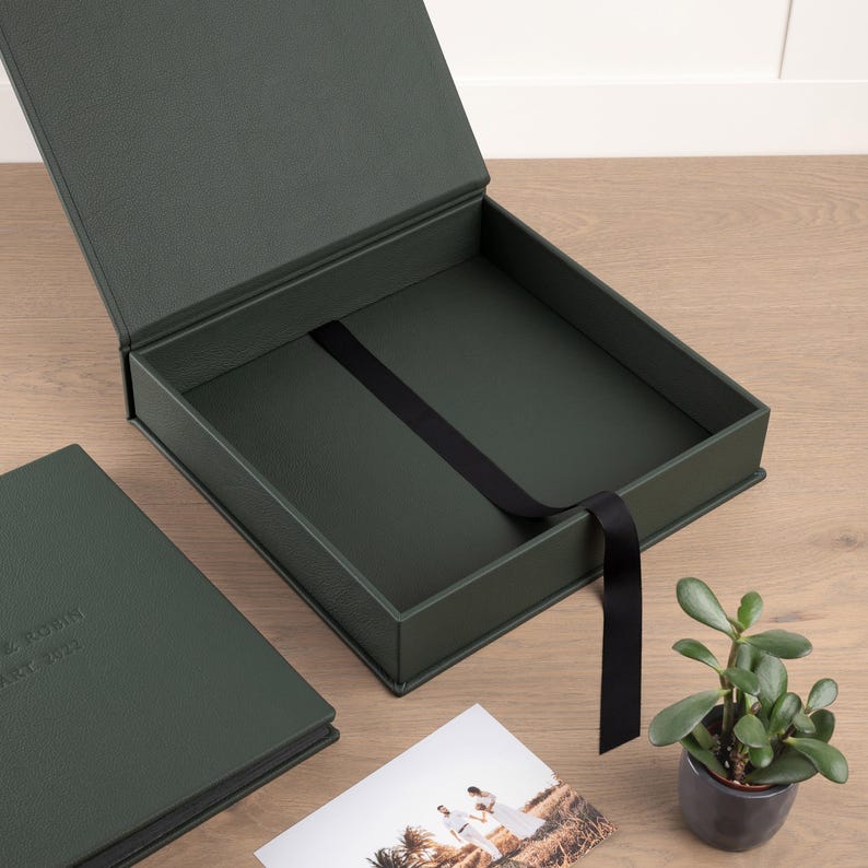 May include: An open, dark green presentation box with a black ribbon, next to a matching album. The box is on a wooden surface, with a small potted plant and a photograph of a couple. The album has text on the cover.