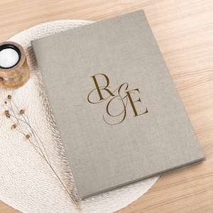 Puede incluir: Un álbum de fotos beige cubierto de lino con las iniciales "R & E" en marrón. El álbum está sobre un salvamanteles blanco tejido. Un portavelas de madera y ramitas secas a la izquierda. El álbum está cerrado.