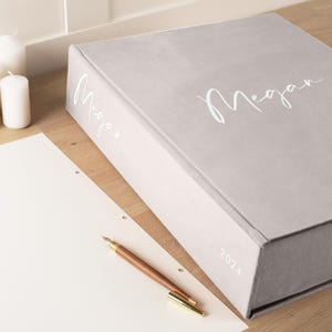 K&ouml;nnte beinhalten: Ein hellgraues, rechteckiges Notizbuch mit dem Namen "Megan" in wei&szlig;er Schrift. Das Jahr "2024" ist auf dem Buchr&uuml;cken aufgedruckt. Ein Holzstift mit goldfarbener Spitze liegt auf einem wei&szlig;en Blatt Papier. Zwei wei&szlig;e Kerzen sind im Hintergrund.