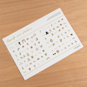 Pode incluir: Uma folha de autocolantes com tema de casamento. Os autocolantes apresentam v&aacute;rias ilustra&ccedil;&otilde;es, incluindo uma noiva e um noivo, cora&ccedil;&otilde;es, ta&ccedil;as de champanhe e as palavras "happily ever stickers". A folha tem detalhes e texto dourados.