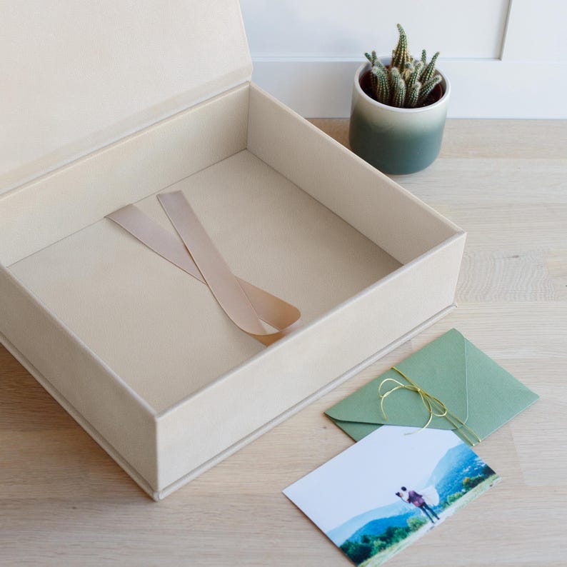 May include: A large, open, square cream-coloured box with a matching ribbon inside. Beside the box sits a small green potted cactus, a sage green envelope tied with thin gold string, and a rectangular photograph showing a person in a landscape.