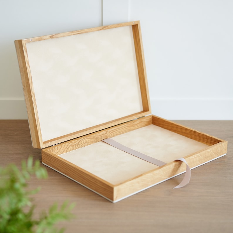 May include: Open wooden box with a light-colored interior and a matching lid. The box has a light brown wood frame with a white border. A light gray ribbon is inside the box.