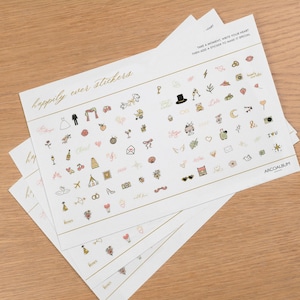 Pode incluir: V&aacute;rias folhas de autocolantes brancos com texto dourado que diz "happily ever stickers". Os autocolantes apresentam pequenas ilustra&ccedil;&otilde;es coloridas de artigos relacionados com casamentos, como um vestido de noiva, an&eacute;is e ta&ccedil;as de champanhe. O texto "Take a moment, write your heart then add a sticker to make it special" tamb&eacute;m &eacute; vis&iacute;vel.