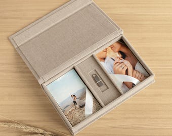 May include: A beige linen-covered keepsake box with a hinged lid, featuring two rectangular compartments for photographs and a central slot containing a small metallic USB flash drive. The box holds two printed photos of a couple in an outdoor setting.
