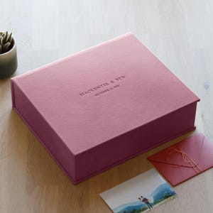 Könnte beinhalten: Eine staubrosa Präsentationsbox mit dem geprägten Text "MacKenzie & Ben October 21, 2023." Ein Foto und ein passender Umschlag mit einer Schnur sind neben der Box. Eine kleine Sukkulentenpflanze befindet sich im Hintergrund.