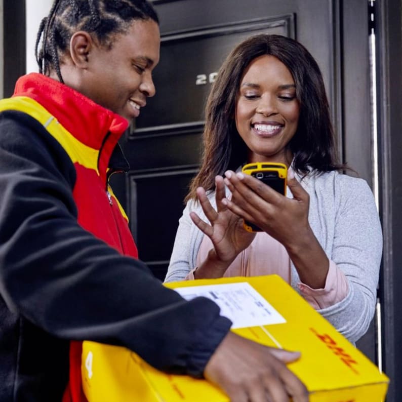 May include: A delivery person in a yellow and red uniform hands a yellow package with the DHL logo to a woman holding a smartphone. The woman is smiling and looking at the phone.