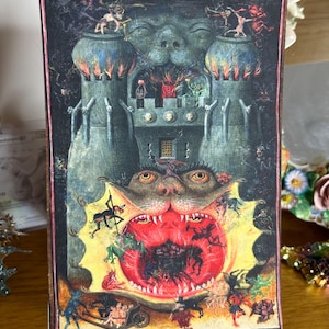 Puede incluir: Impresión enmarcada que presenta una ilustración gótica surrealista. La obra de arte representa un castillo con figuras demoníacas, una gran boca abierta y una escena infernal y ardiente. La paleta de colores incluye azules oscuros, rojos y verdes.