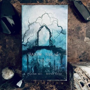 Pode incluir: Um baralho de cartas de tarô retangular intitulado "Shadow Journey Tarot". A capa apresenta uma ilustração em aquarela azul e cinza de um arco e árvores. Os nomes J. Edward Neill e Heather Kelter também são visíveis.