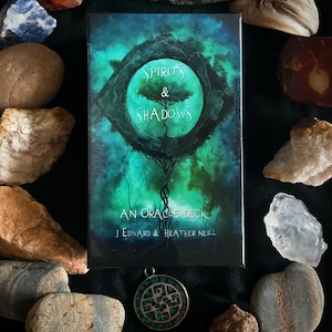 May include: An oracle deck box with the title "SPIRITS & SHADOWS" in white text, featuring a tree design within a circular frame. The box is surrounded by rocks and a pendant. The box is rectangular.