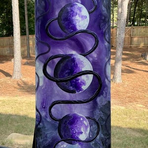 May include: A vertical rectangular canvas painting featuring a long black snake winding through three circular moon-like spheres. The background is a deep, swirling purple and black nebula-like wash, creating a dark, celestial atmosphere.