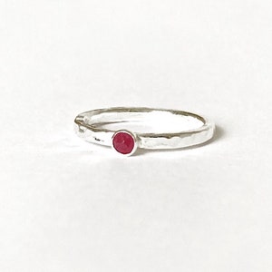 May include: A silver ring with a hammered finish, featuring a small, round, red gemstone. The ring's design is simple, set against a white backdrop, emphasizing the gemstone's colour and the ring's texture.