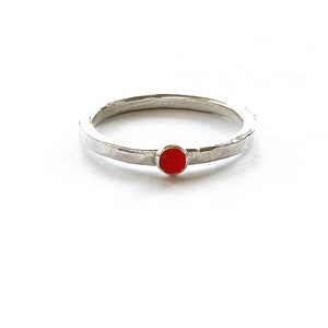 May include: A silver ring with a hammered texture and a small, round, red gemstone. The ring is set against a white background. The gemstone is approximately 0.25 cm in diameter, adding a pop of colour to the minimalist design.