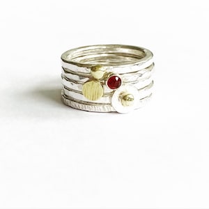 May include: A silver ring set with five bands, each with a textured finish. The ring features three gold accents, including a round bead, a flat disc, and a larger disc with a raised centre. A red gemstone is set in the centre of the ring.